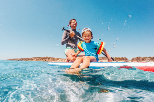 Kind mit bunten Schwimmfl&uuml;geln auf Paddelboard mit Vater auf dem Meer