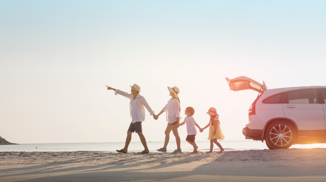 Vater, Mutter und zwei Kinder halten sich an den Händen und laufen in einer Reihe am Strand entlang nach links, Vater zeigt die Richtung mit seinem Arm und rechts von ihnen steht ihr Auto am Strand